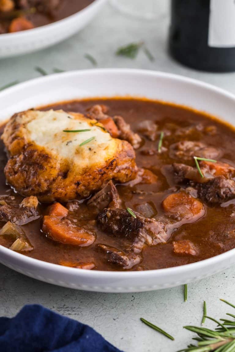 side shot of a white ceramic soup bowl filled with a tomato based beef stew with large chunks of beef, and carrots visible. A large rosemary dumpling is sitting in the upper left area of the bowl. The bottom edge of a bottle of red wine is visible in the upper right corner of the picture, and another bowl of stew is peeking-in in the upper left corner of the picture.