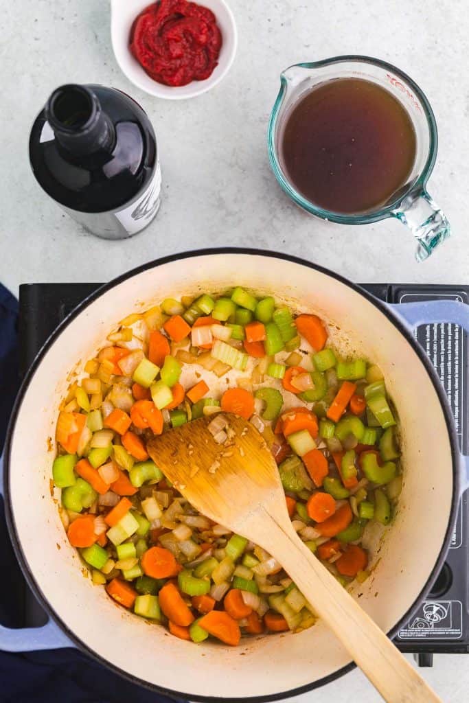 Overhead shot of a Dutch oven with carrots, onions, and celery sauteing. Above the Dutch oven in the photo is a bottle of red wine, a small bowl of tomato paste, and a measuring cup of beef stock, all to be added next.