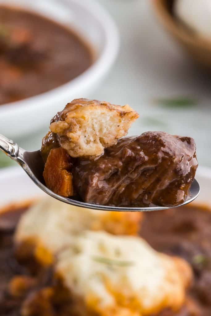 Close up side view of a spoon holding a large chunk of tender slow cooked beef, some carrot, and a piece of the rosemary dumpling. In the background there is a large bowl of the beef stew and dumplings.