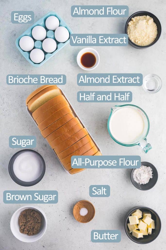 Overhead shot of all the ingredients used in make Brioche French Toast Casserole. Ingredients include: eggs, almond flour, all-purpose flour, vanilla extract, almond extract, brioche bread, half and half, sugar, brown sugar, salt, and butter.