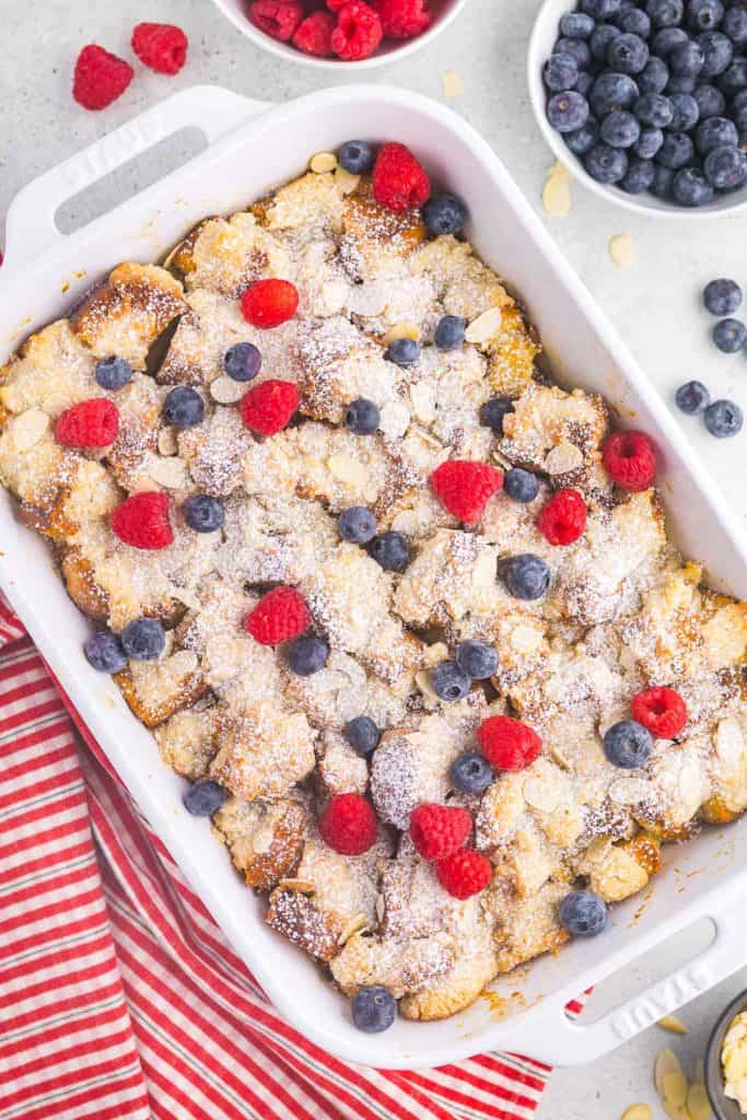 Overhead shot of a white casserole dish filled with golden brown brioche french toast casserole, sprinkled with icing sugar and dotted with some fresh blueberries and raspberries. The bottom left corner has a red and white striped linen and the upper right corner has two bowls one filled with blueberries and one with raspberries.