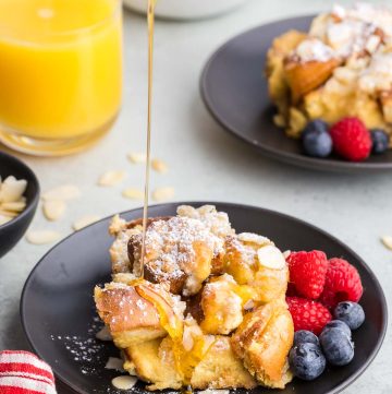 side shot of maple syrup being poured out of a white ceramic jug onto a slice of brioche French toast casserole on a black plate. The golden syrup is glistening in the light and dripping down the front edge of the casserole. Fresh raspberries and blueberries are garnished on the side of the plate, and in the background are other breakfast items like a glass of orange juice and another serving of French toast.