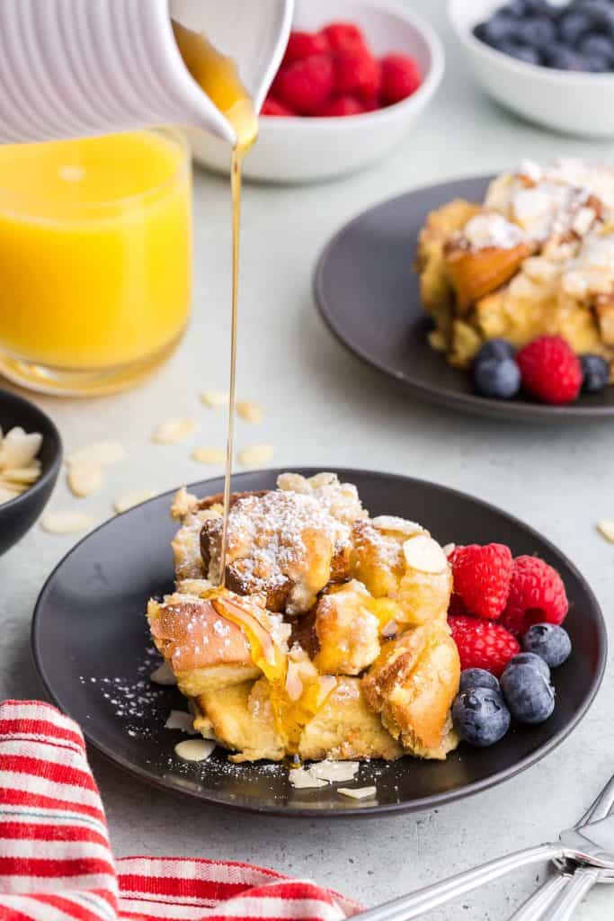 side shot of maple syrup being poured out of a white ceramic jug onto a slice of brioche French toast casserole on a black plate. The golden syrup is glistening in the light and dripping down the front edge of the casserole. Fresh raspberries and blueberries are garnished on the side of the plate, and in the background are other breakfast items like a glass of orange juice and another serving of French toast.