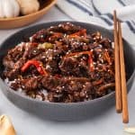 Side shot of a shallow gray ceramic bowl filled with rice and crispy ginger beef in a sticky dark sauce and garnished with sesame seeds. The bottom left corner has some fortune cookies peeking in.
