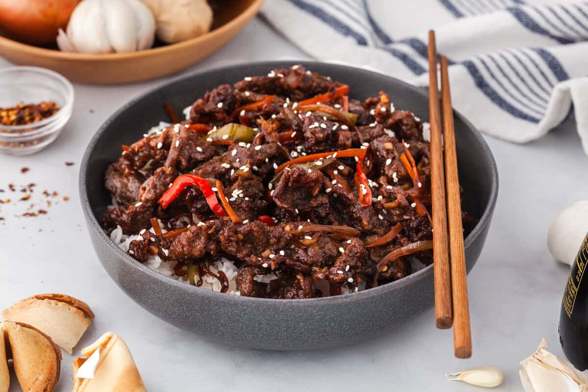 Side shot of a shallow gray ceramic bowl filled with rice and crispy ginger beef in a sticky dark sauce and garnished with sesame seeds. The bottom left corner has some fortune cookies peeking in.