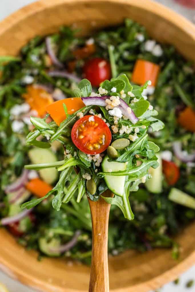 Overhead shot close up of a wooden serving spoon filled with quinoa salad with arugula. On the spoon there is plenty of green leafy arugula was well as a juicy halved cherry tomato, orange bell pepper chunk, some feta cheese, and lots of quinoa. In the background is the serving bowl of salad but it is out of focus.