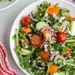 Overhead shot of a white shallow bowl filled with green arugula, red halved cherry tomatoes, orange pepper chunks, cucumber chunks, thin slices of red onion, and quinoa. The bowl is centered vertically but slightly to the right, so the far right of the bowl is cut off. The upper left corner has another bowl of quinoa arugula salad peeking in. The upper right corner has a vine of cheery tomatoes peeking in. The bottom right corner is part of a small black bowl of feta, and the bottom left corner has a red and white striped linen present.