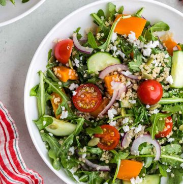 Overhead shot of a white shallow bowl filled with green arugula, red halved cherry tomatoes, orange pepper chunks, cucumber chunks, thin slices of red onion, and quinoa. The bowl is centered vertically but slightly to the right, so the far right of the bowl is cut off. The upper left corner has another bowl of quinoa arugula salad peeking in. The upper right corner has a vine of cheery tomatoes peeking in. The bottom right corner is part of a small black bowl of feta, and the bottom left corner has a red and white striped linen present.