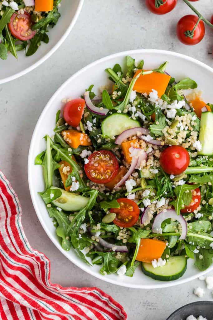 Overhead shot of a white shallow bowl filled with green arugula, red halved cherry tomatoes, orange pepper chunks, cucumber chunks, thin slices of red onion, and quinoa. The bowl is centered vertically but slightly to the right, so the far right of the bowl is cut off. The upper left corner has another bowl of quinoa arugula salad peeking in. The upper right corner has a vine of cheery tomatoes peeking in. The bottom right corner is part of a small black bowl of feta, and the bottom left corner has a red and white striped linen present.