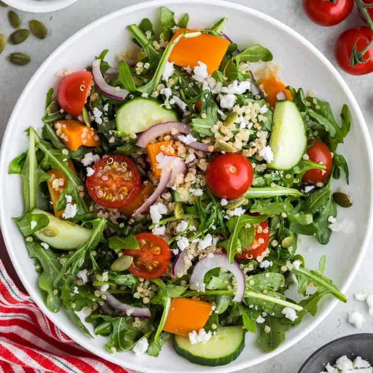 Overhead close up shot of a white shallow bowl filled with green arugula, red halved cherry tomatoes, orange pepper chunks, cucumber chunks, thin slices of red onion, and quinoa. The bowl is centered and takes up the entire photo.