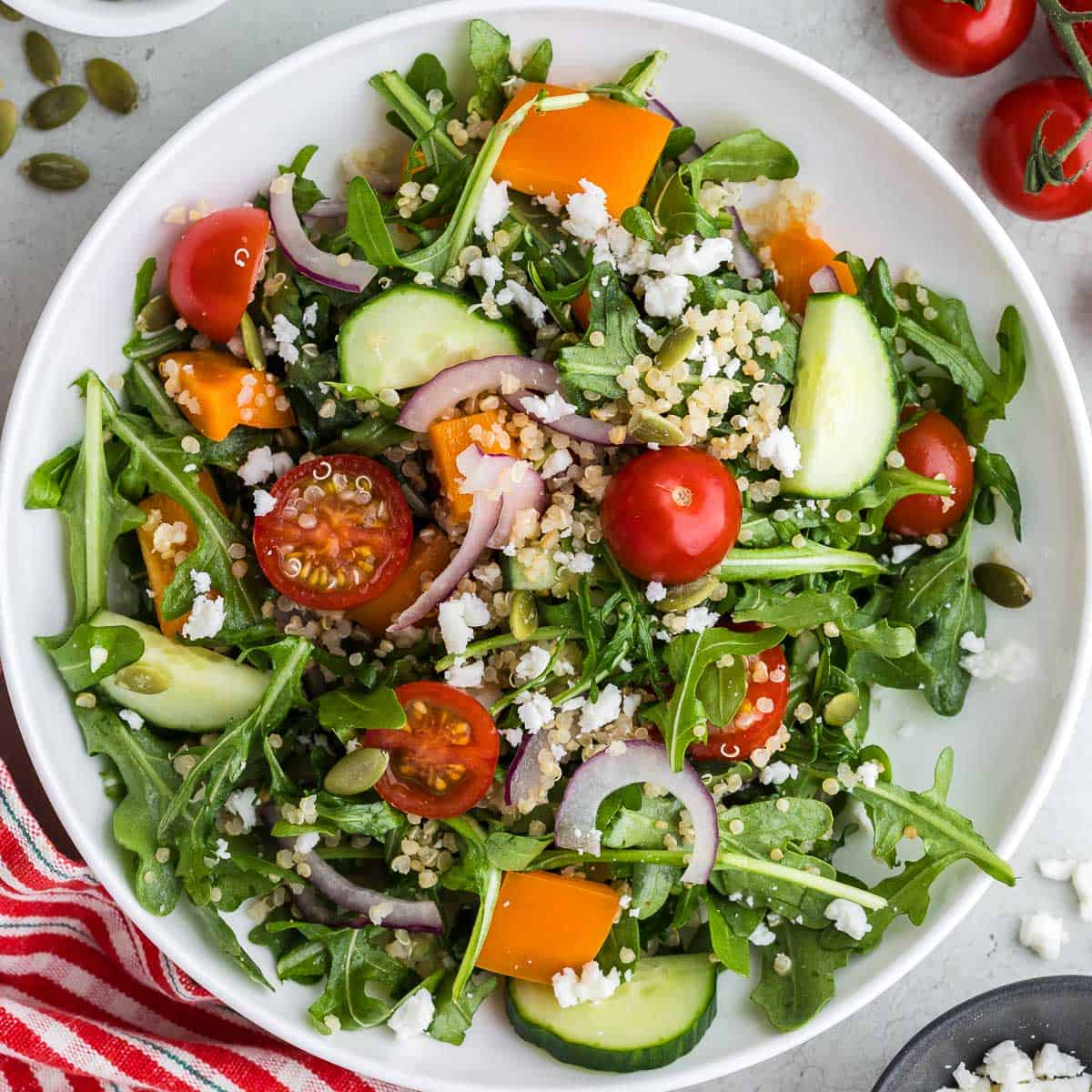 Overhead close up shot of a white shallow bowl filled with green arugula, red halved cherry tomatoes, orange pepper chunks, cucumber chunks, thin slices of red onion, and quinoa. The bowl is centered and takes up the entire photo.