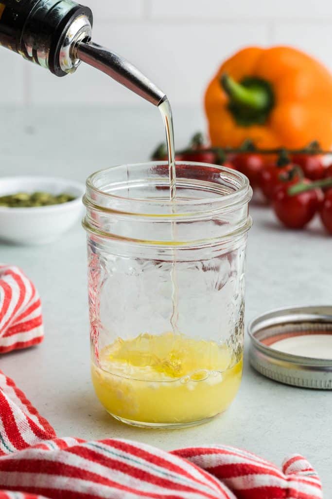 Side shot of a small mason jar filled with fresh lemon juice, dijon mustard, honey and garlic. A spout is coming in from the upper left corner and olive oil is being poured out of it and into the mason jar. In the background and slightly out of focus is an orange pepper and some cherry tomatoes.