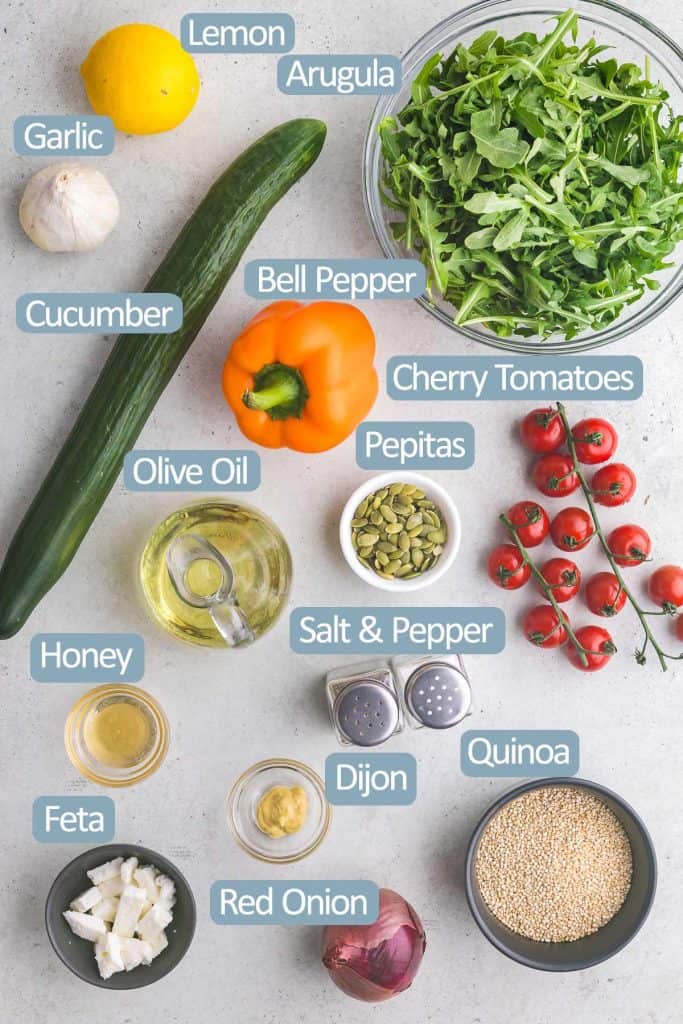 Overhead shot of the all the ingredients needed to make this quinoa salad with arugula. Ingredients include: lemon, garlic, arugula, cucumber, bell pepper, cherry tomatoes, pepitas, olive oil, salt and pepper, honey, dijon mustard, feta cheese, red onion, and quinoa.