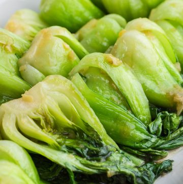 Close up shot of vibrant green cooked Shanghai bok choy organized in the same direction on an elongated white serving platter. They are all covered in a translucent sauce with small chunks of ginger dispersed throughout.