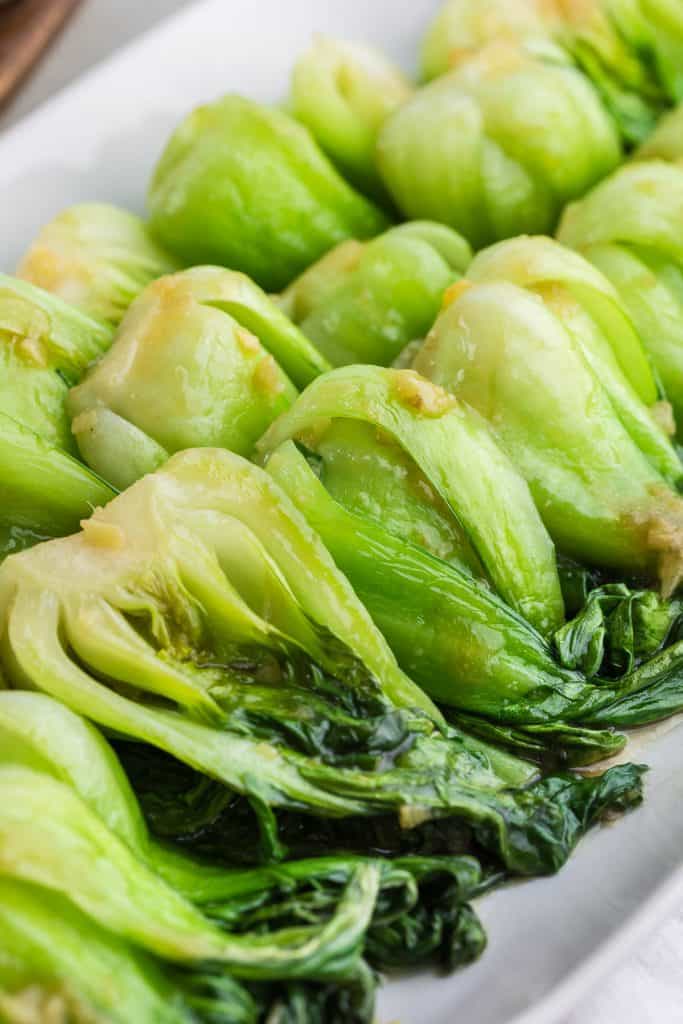 Close up shot of vibrant green cooked Shanghai bok choy organized in the same direction on an elongated white serving platter. They are all covered in a translucent sauce with small chunks of ginger dispersed throughout.