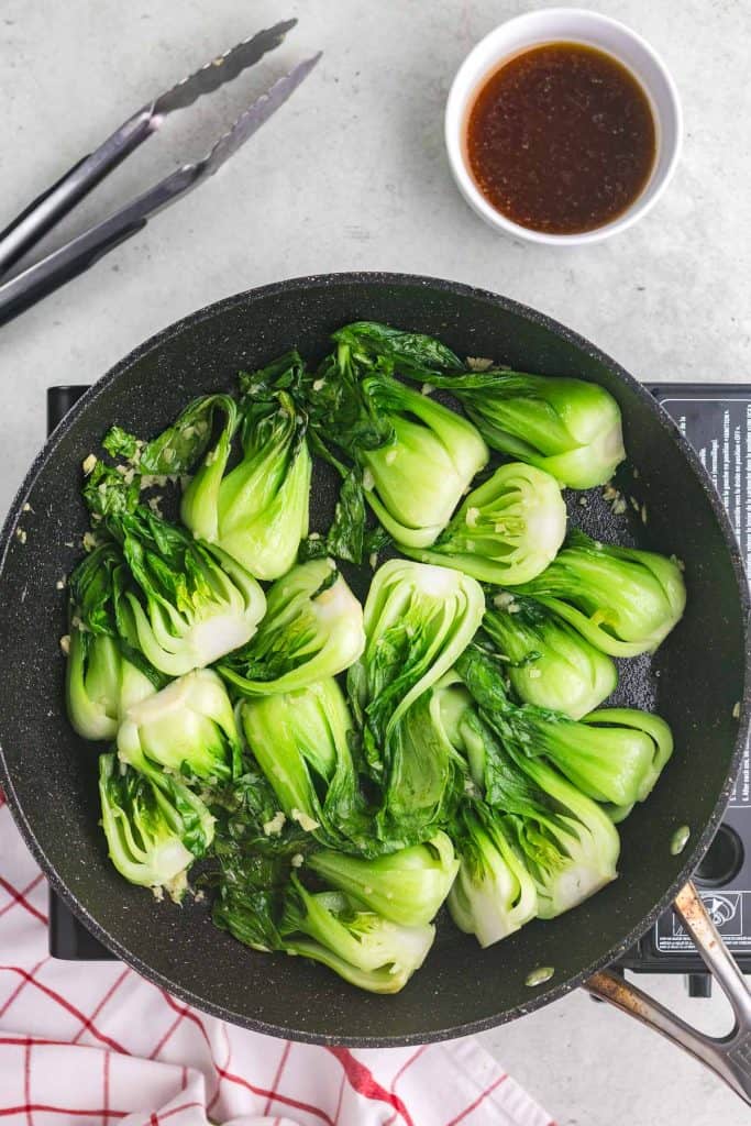 Overhead shot of a black frypan filled with vibrant green shanghai bok choys all stir fried until soft.  Near the top right is a small white bowl with a brown sauce which will be added next, and in the top left corner are tongs which have been used to toss the Shanghai bok choy while cooking.