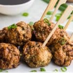 Close up shot of Air Fryer Chicken Meatballs on a white rectangular serving plate. Wooden bamboo skewers are poking out of four of the meatballs, ready to be picked up and dipped into the Asian sauce in a small white bowl in the background. Green onions are scattered on and around the meatballs