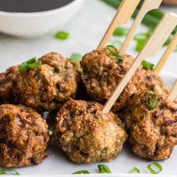 Close up shot of Air Fryer Chicken Meatballs on a white rectangular serving plate. Wooden bamboo skewers are poking out of four of the meatballs, ready to be picked up and dipped into the Asian sauce in a small white bowl in the background. Green onions are scattered on and around the meatballs