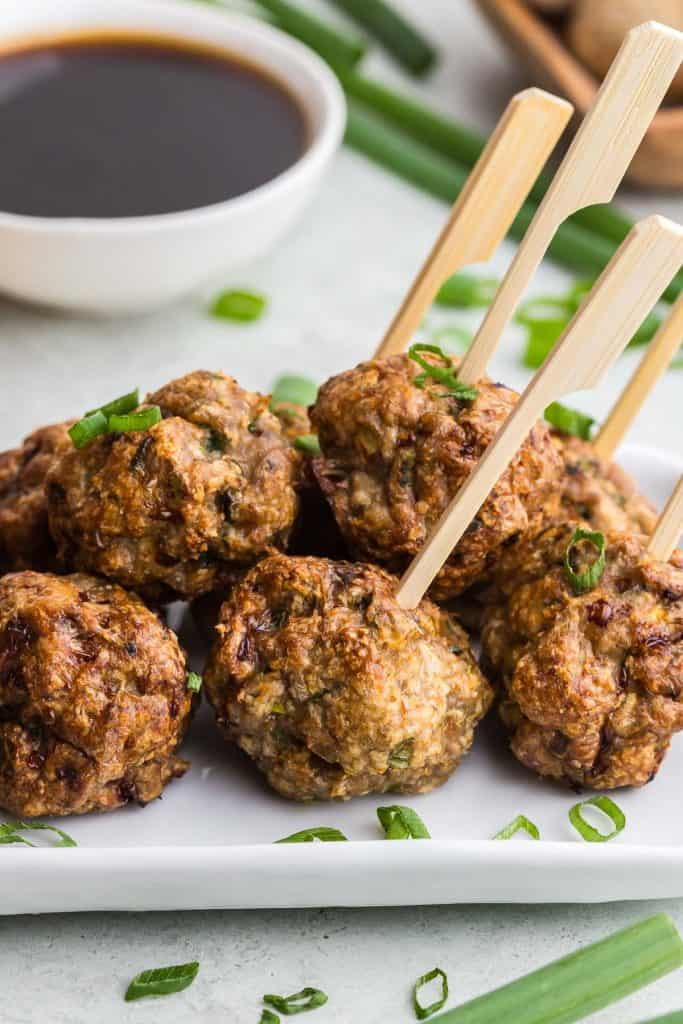Close up shot of Air Fryer Chicken Meatballs on a white rectangular serving plate.  Wooden bamboo skewers are poking out of four of the meatballs, ready to be picked up and dipped into the Asian sauce in a small white bowl in the background.  Green onions are scattered on and around the meatballs
