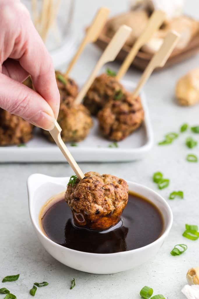 Side shot of a small white bowl filled with a dark Asian inspired sauce.  An air fryer chicken meatball is being dipped into the sauce.