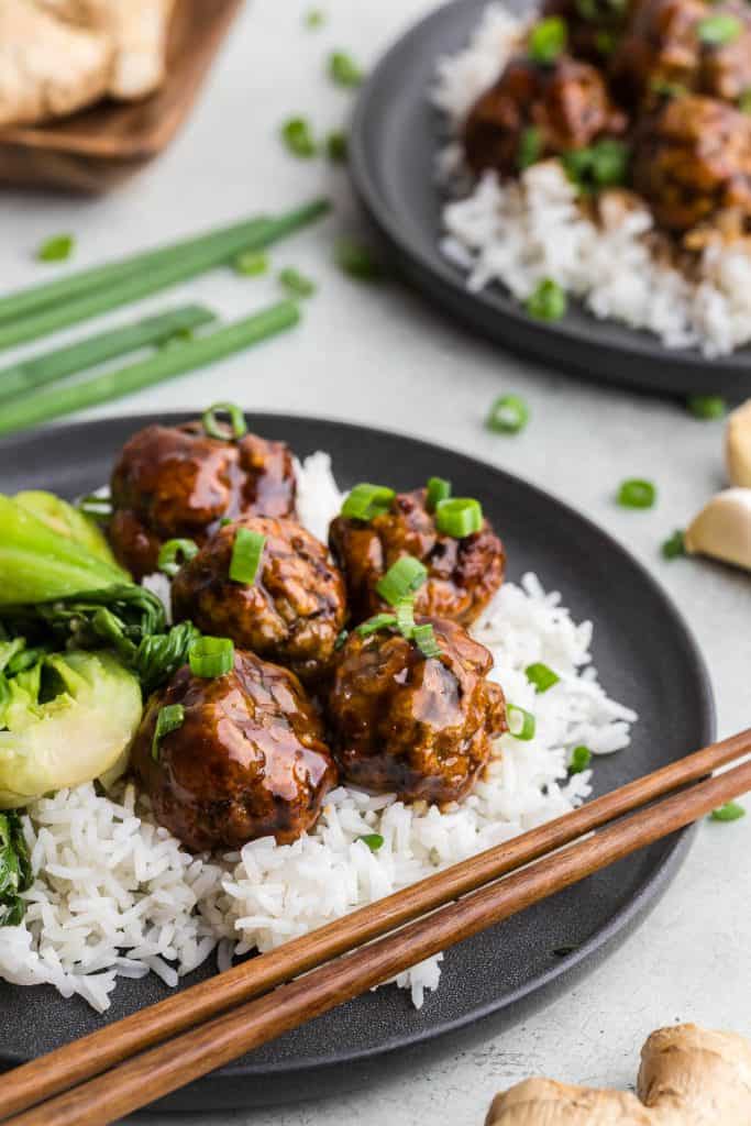 Side shot of a dark grey ceramic plate filled with rice and topped with 5 air fry chicken meatballs smothered in an Asian inspired sauce. Beside the meatballs are some sauteed Shanghai baby bok choy. Wooden chopsticks are resting on the plate.
