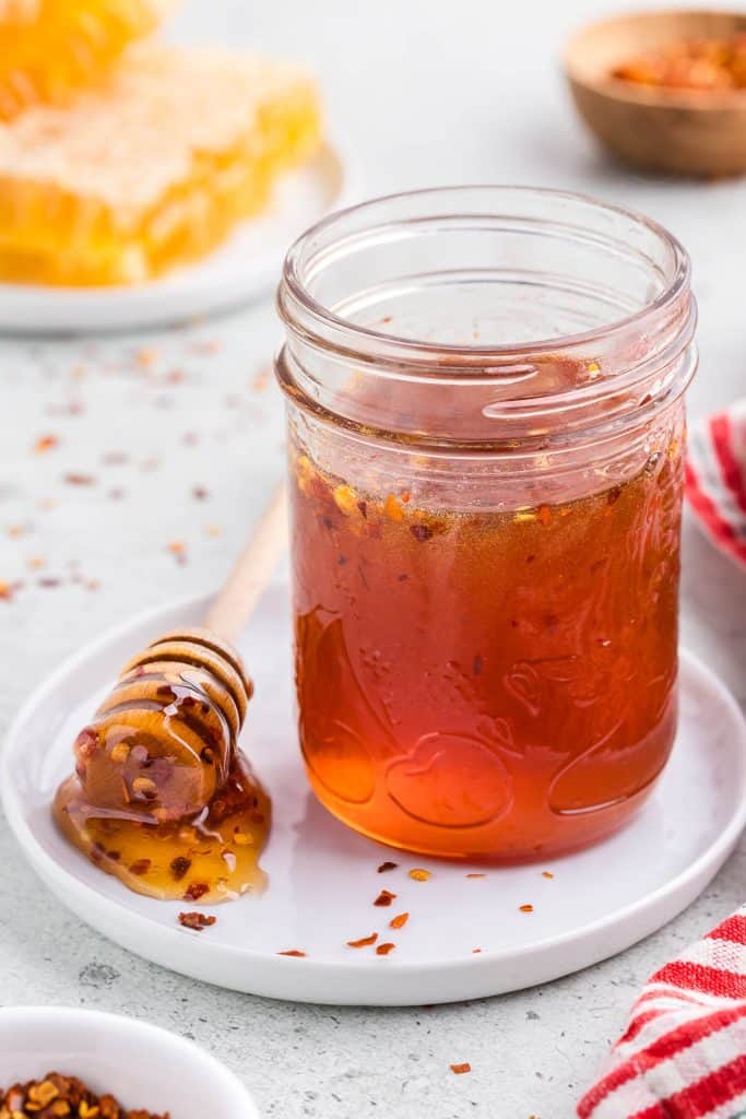 Side shot of a small mason jar filled with golden brown honey with specs of chili flakes floating in it. The jar is on a small white ceramic plate and there is a honey dipper resting on the plate in a small pool of hot hone. In the background there is a small plate of fresh honeycomb and a small wooden pinch bowl of chili pepper flakes.