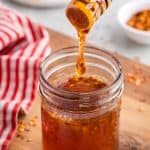Side shot of a small mason jar filled with dark golden brown honey with a few specs of chili pepper flakes in it. A wooden honey dipper has just pulled out of the jar and a large dollop of honey is dripping from the dipper and falling back into the jar. In the background is a white plate with some honeycomb on it, and a small pinch bowl of chili pepper flakes.