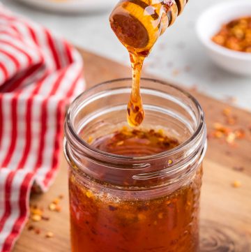 Side shot of a small mason jar filled with dark golden brown honey with a few specs of chili pepper flakes in it. A wooden honey dipper has just pulled out of the jar and a large dollop of honey is dripping from the dipper and falling back into the jar. In the background is a white plate with some honeycomb on it, and a small pinch bowl of chili pepper flakes.