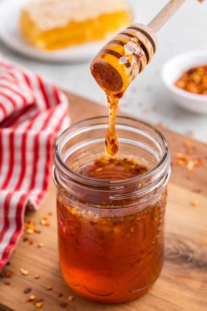 Side shot of a small mason jar filled with dark golden brown honey with a few specs of chili pepper flakes in it. A wooden honey dipper has just pulled out of the jar and a large dollop of honey is dripping from the dipper and falling back into the jar. In the background is a white plate with some honeycomb on it, and a small pinch bowl of chili pepper flakes.