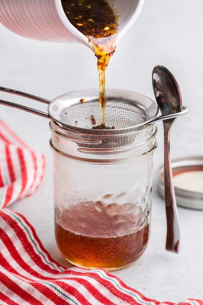 Side shot of a small mason jar with a fine mesh sieve sitting atop. Freshly prepared hot honey is being poured out of a white ceramic jug into the jar through the sieve in order to filter out the chili flakes. The mason jar is 1/4 filled with a dark honey, and the sieve has many chili flakes left behind.