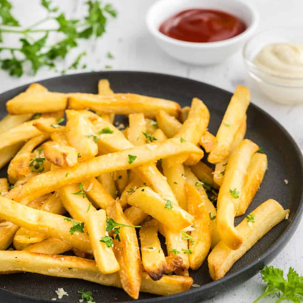 Side shot of a pile of golden parmesian truffle fries garnished with fresh parsley on a dark grey ceramic plate.  In the background are two pinch bowls, one filled with a bright red ketchup and the other with a creamy white garlic aioli.