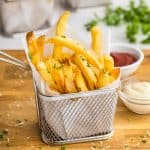 Side shot of golden brown French Fries neatly stacked vertically into a parchment paper lined stainless steel small fry basket. The fry basket is sitting on a large wooden cutting board, and there are two small pinch bowls off to the right, one filled with ketchup and the other with a garlic aioli. In the background there are two more small French fry baskets filled with golden truffle fries.