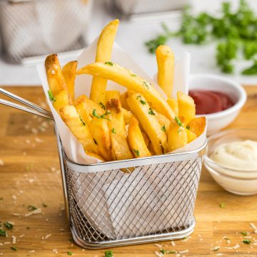 Side shot of golden brown French Fries neatly stacked vertically into a parchment paper lined stainless steel small fry basket. The fry basket is sitting on a large wooden cutting board, and there are two small pinch bowls off to the right, one filled with ketchup and the other with a garlic aioli. In the background there are two more small French fry baskets filled with golden truffle fries.