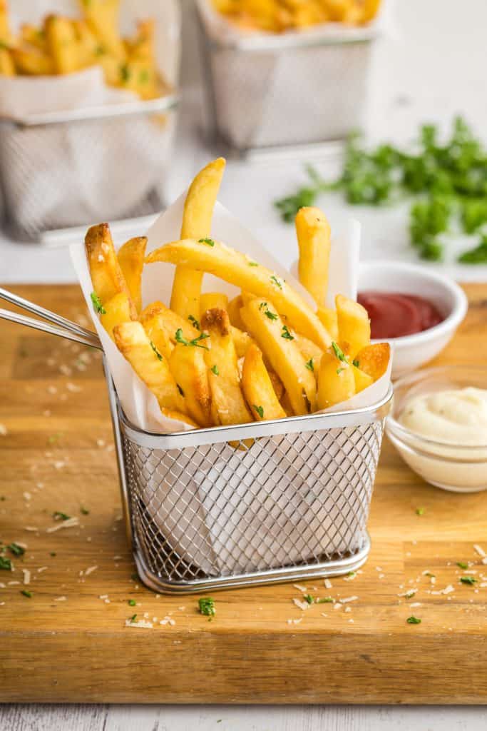 Side shot of golden brown truffle fries neatly stacked vertically into a parchment paper lined stainless steel small fry basket.  The fries are garnished with shredded parmesan and parsley flakes.  The fry basket is sitting on a large wooden cutting board, and there are two small pinch bowls off to the right, one filled with ketchup and the other with a garlic aioli.  In the background there are two more small French fry baskets filled with golden truffle fries.