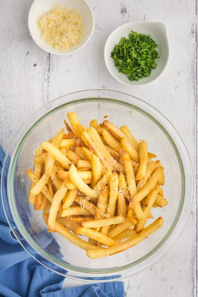 Overhead shot of a large glass bowl filled with golden French fries.  Shredded parmesan has been lightly added to the fries along with the truffle oil.  Above the bowl are two small pinch bowls, one filled with freshly grated parmesan and the other with fresh chopped parsley.