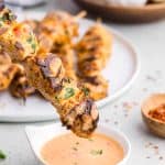 close up side shot of a skewer of chicken with black grill marks and garnished with fresh chopped parsley is just about to be dipped into a pale orange bang bang sauce in a small white bowl. In the background is a plate full of more chicken skewers and a small wooden bowl with fresh onion and garlic.