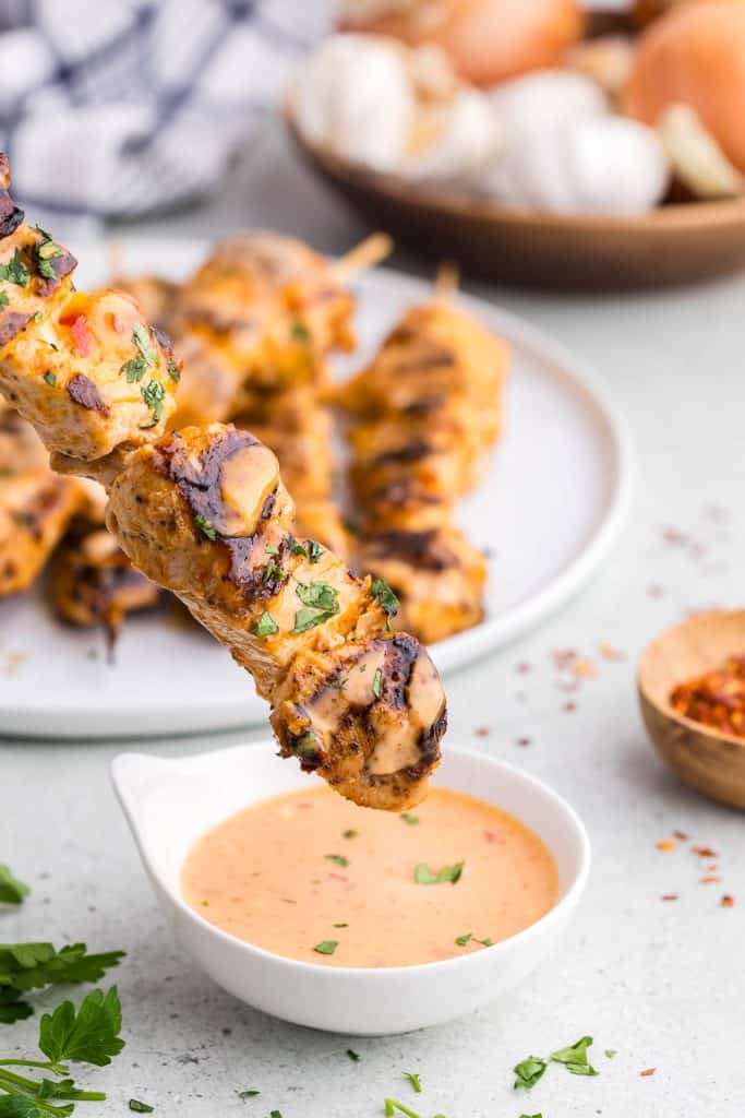 close up side shot of a skewer of chicken with black grill marks and garnished with fresh chopped parsley is just about to be dipped into a pale orange bang bang sauce in a small white bowl. In the background is a plate full of more chicken skewers and a small wooden bowl with fresh onion and garlic.