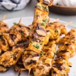 Side shot of a plate filled with 8 skewers of grilled bang bang chicken skewers. Each skewer has black grill marks, is drizzled with a pale orange colored bang bang sauce, and garnished with parsley.