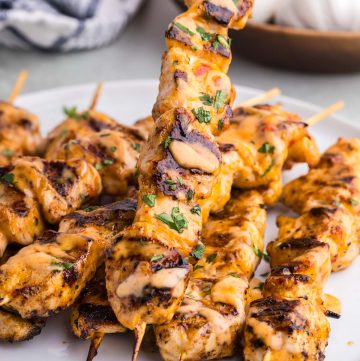 Side shot of a plate filled with 8 skewers of grilled bang bang chicken skewers. Each skewer has black grill marks, is drizzled with a pale orange colored bang bang sauce, and garnished with parsley.