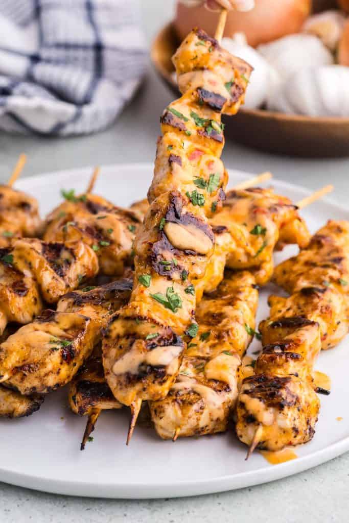 Side shot of a plate filled with 8 skewers of grilled bang bang chicken skewers. Each skewer has black grill marks, is drizzled with a pale orange colored bang bang sauce, and garnished with parsley.