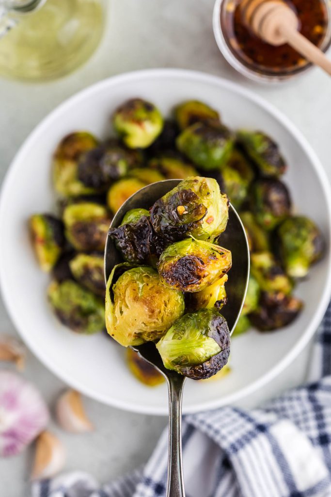Overhead shot of a white ceramic bowl filled with dark green roasted brussels sprouts with tender yellow centers. A silver metal serving spoon is lifting a serving of hot honey Brussels sprouts out of the bowl towards the camera lens.