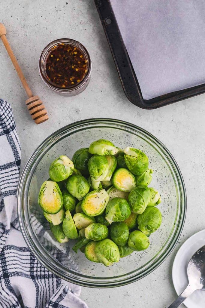 Overhead shot of a large glass mixing bowl in the lower 2/3rds of the frame. The bowl is filled with bright light green halved Brussels sprouts that have been seasoned with garlic powder, onion powder, salt, pepper, and olive oil. Above the bowl on the left is a small jar of dark hot honey, and on the right is the corner of a baking pan lined with parchment paper