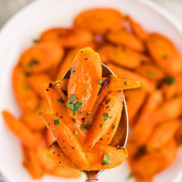 Overhead shot of a serving spoon of hot honey carrots being pulled up out of the bowl towards the camera lens. The bright orange carrots are cut on the diagonal into large 1/2" chunks and are garnished with chopped parsley.