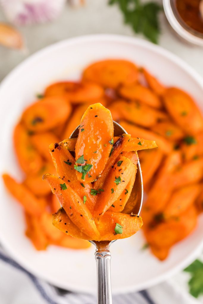 Overhead shot of a serving spoon of hot honey carrots being pulled up out of the bowl towards the camera lens. The bright orange carrots are cut on the diagonal into large 1/2" chunks and are garnished with chopped parsley.