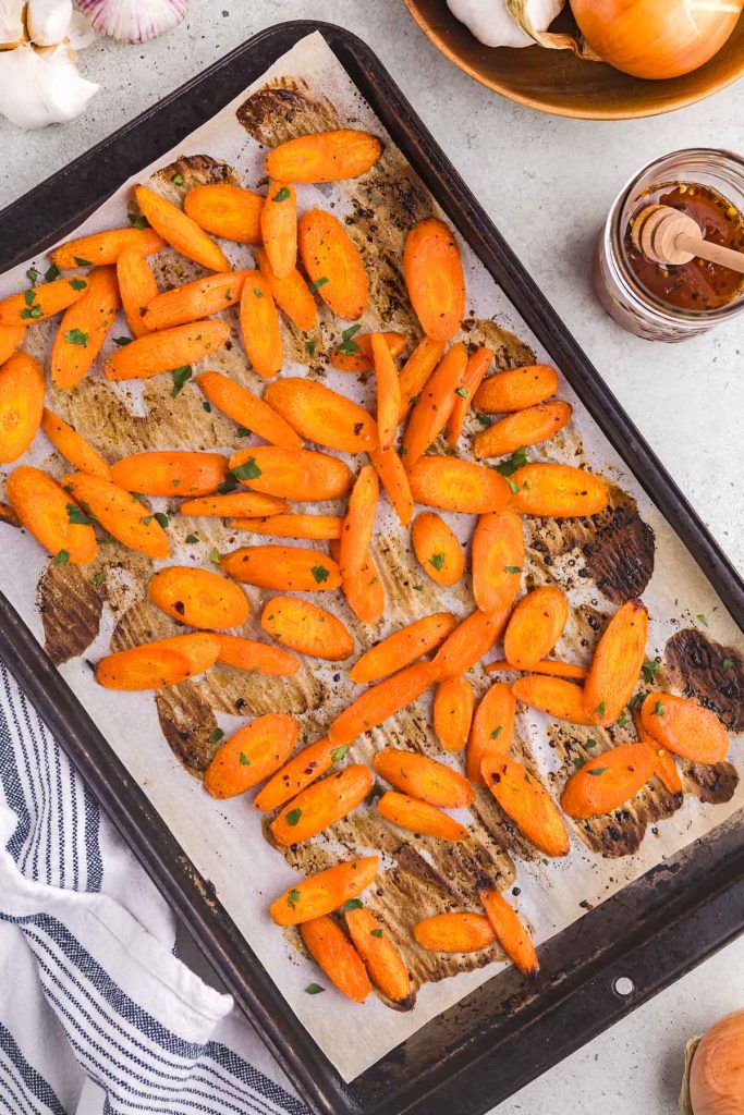 Overhead shot of the hot honey carrots on a baking sheet coming out of the oven after being roasted. The carrots are bright orange with small areas of charring from being roasted. In the upper right corner there is a wooden bowl with some fresh garlic bulbs and onions peeking in. Also there is a small jar of hot honey with a honey dipper resting in it, next to the bowl of garlic and onions.