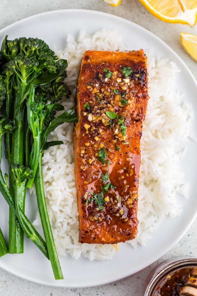 Overhead shot of a a white plate with hot honey salmon served over white rice and a side of broccolini. The hot honey salmon is a strong orange color with a golden brown sear on it and finished off with a shiny hot honey glaze.