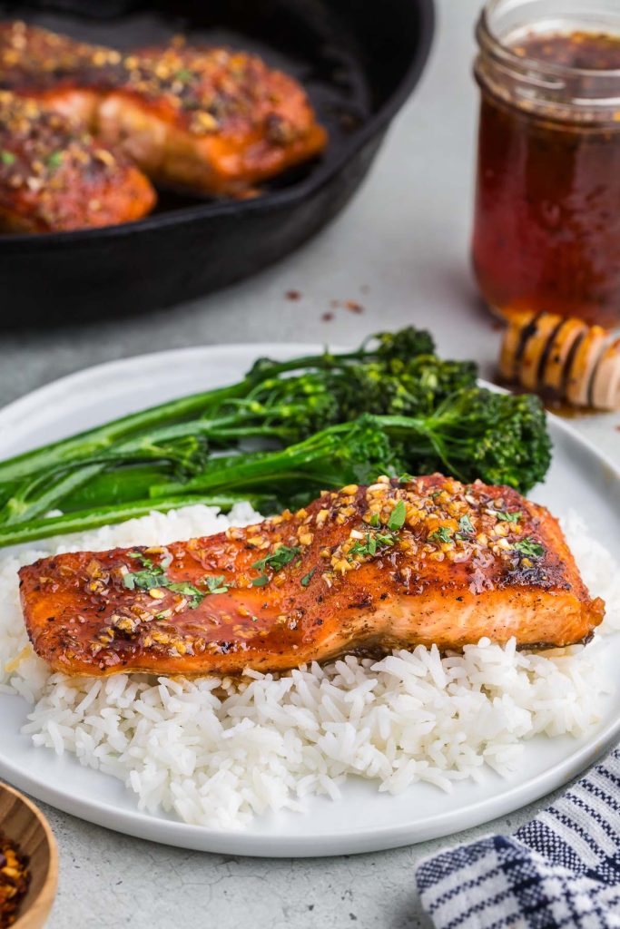 A bright orange hot honey salmon fillet on a bed of white rice, with bright green broccolini served on the side.  In the background on the left is a black cast iron pan with more salmon in it ready to be served.  In the background on the right is a small jar of hot honey with a honey dipper resting next to it.