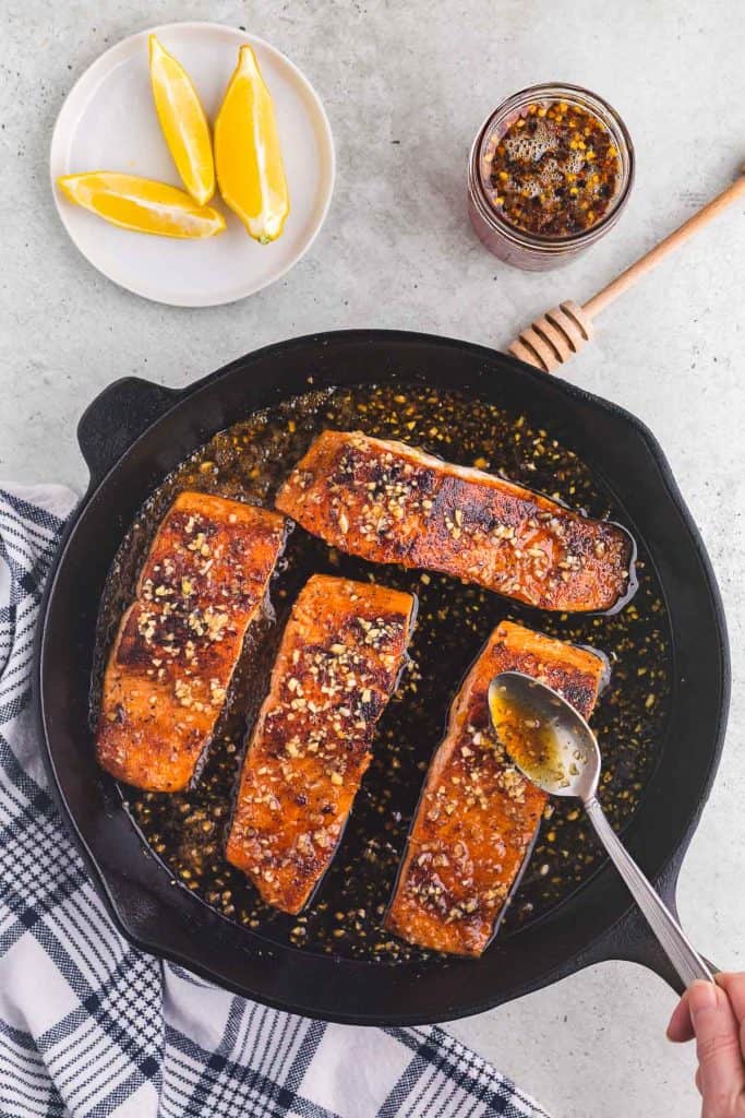 Overhead shot of four pieces of bright orange salmon cooking in a black cast iron skillet. A silver spoon is pouring the hot honey glaze over one of the fillets. All the fillets are glistening with the hot honey sauce and small chunks of garlic are distributed throughout the glaze. Outside the cast iron skillet is a small white plate with lemon wedges ready to be served with the salmon, as well as a jar of hot honey.