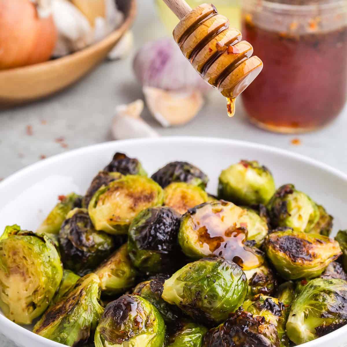 Side shot of a white ceramic bowl filled with dark green roasted hot honey Brussels sprouts. A honey dipper is hovering over the serving dish with a drip of hot honey just about to drip down onto the sprouts. In the background is a glass jar filled with amber colored hot honey, and a shallow wooden bowl with onions and garlic bulbs.