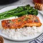 Side shot of a fillet of hot honey salmon sitting atop of a bed of white rice with a side of broccolini all on a white ceramic plate. The hot honey salmon has a golden crispy sear on the flesh with small chunks of garlic and a golden brown hot honey glaze, garnished with fresh parsley.